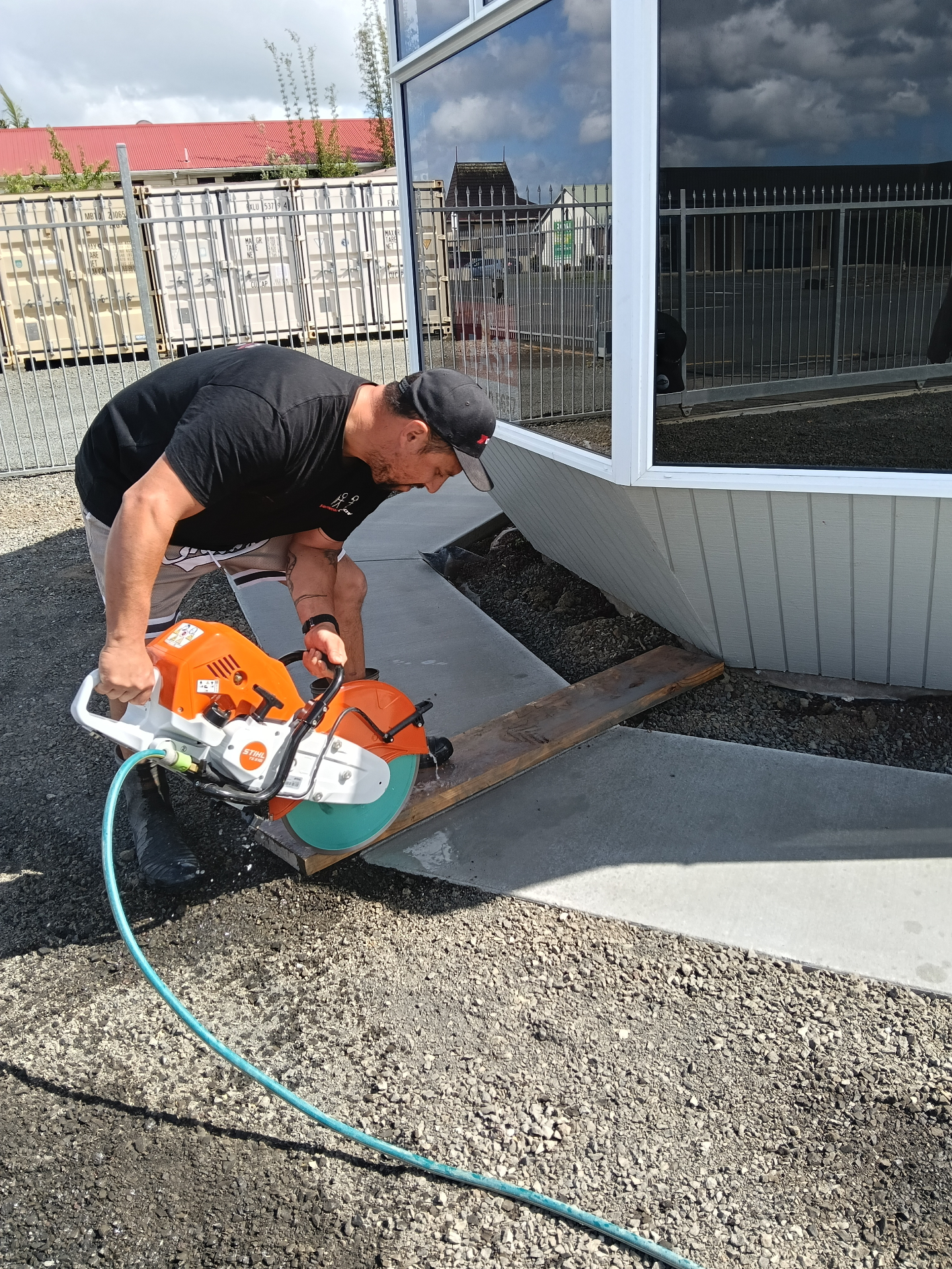 Worker using an orange and white Stihl concrete saw to cut a freshly poured concrete slab beside a modern building, by Henwood Concrete