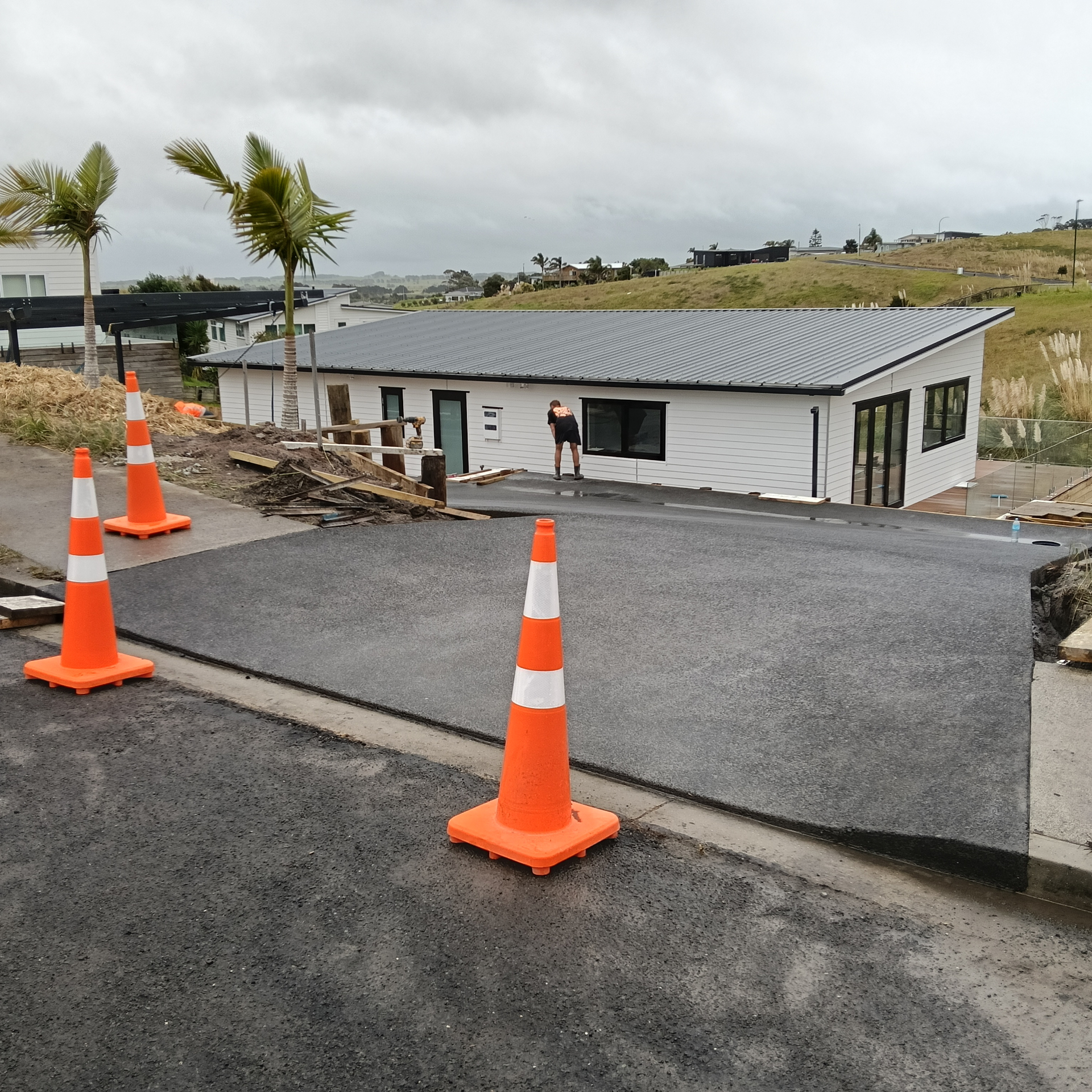 New concrete vehicle crossing on a sloping driveway in front of a modern white house, with traffic cones and a worker finishing the surface by Henwood Concrete