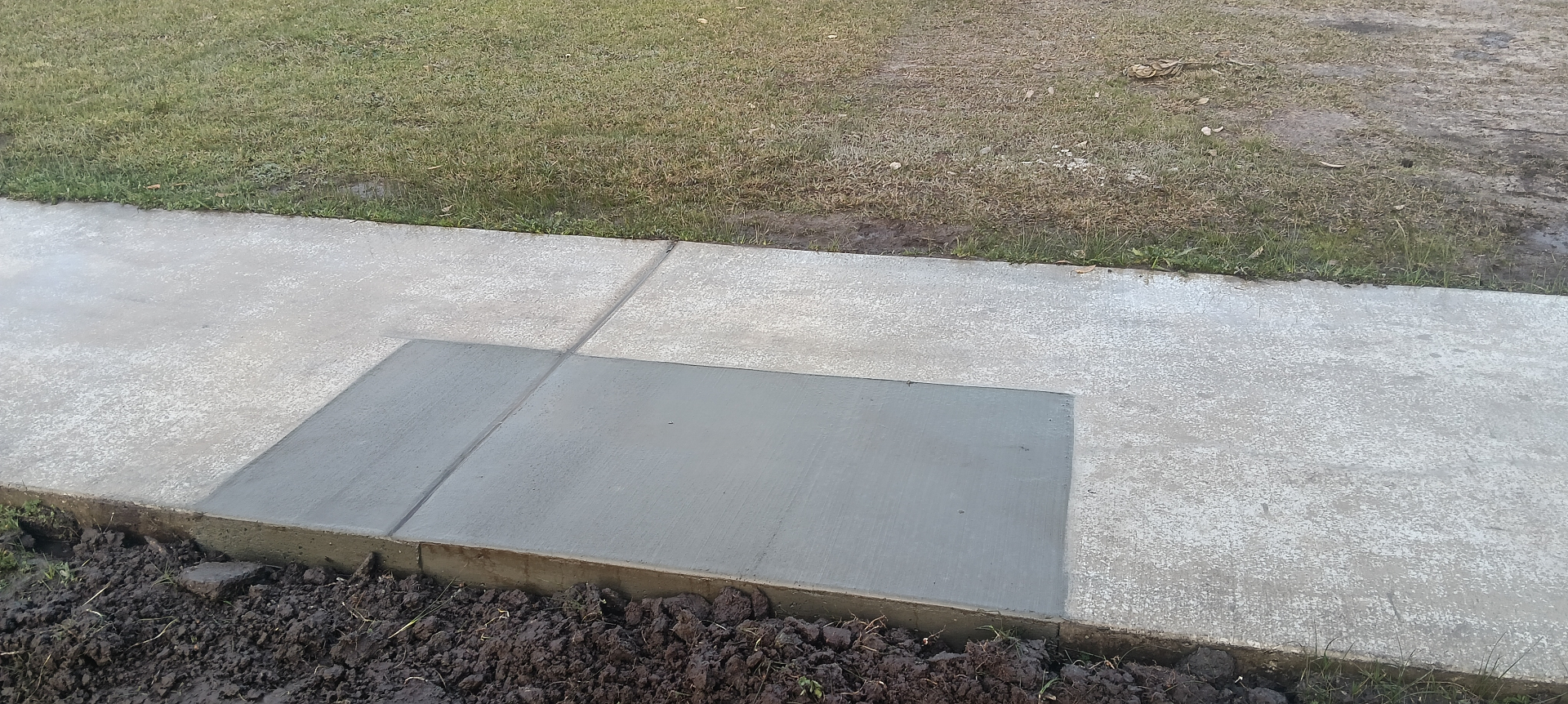 Two dark-grey concrete repair sections set into an existing lighter concrete footpath, by Henwood Concrete.