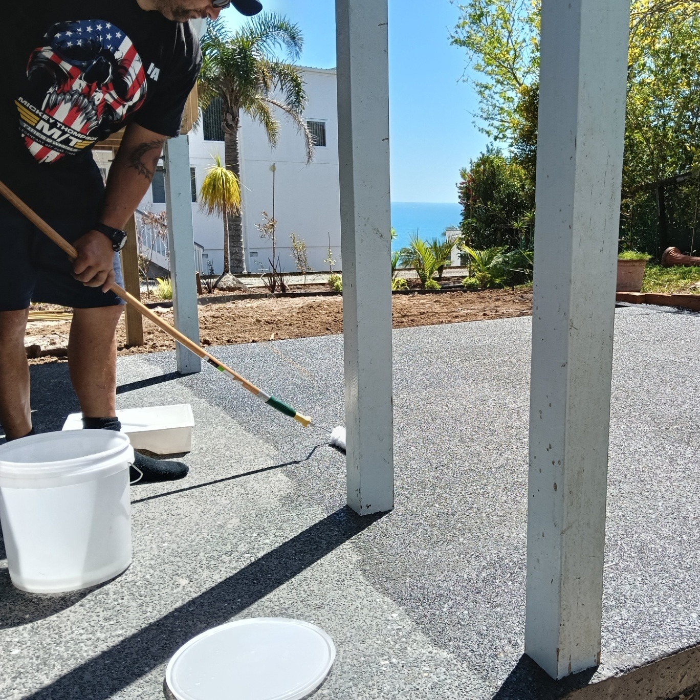 Worker applying sealer to an exposed aggregate concrete patio with the ocean in the background, by Henwood Concrete.