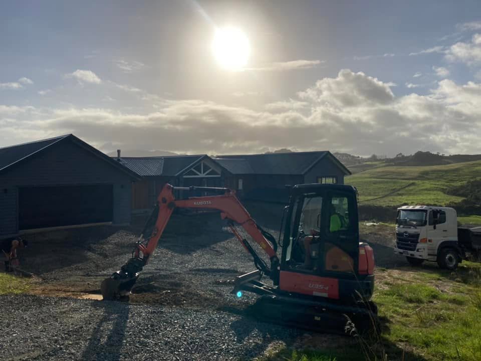 Kubota excavator working on a prepared driveway basecourse area in front of a modern home, with a truck nearby and the sun shining, by Henwood Concrete